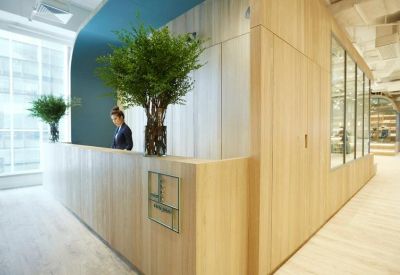 Reception desk with light wood paneling and large leafy plants.