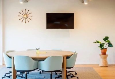 Minimalist meeting room with a round wooden table, light blue chairs, and a wall-mounted screen.