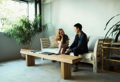 Communal lounge area with a wooden coffee table, indoor plants, and people seated on a white sofa.