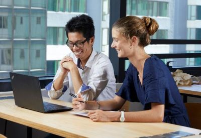 Two colleagues collaborating at a wooden desk with a laptop near a large window.
