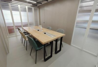 Long wooden collaboration desk with mixed-color chairs in a bright, partitioned office space.