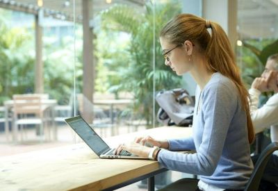 Person working on a laptop at a shared wooden desk with lush greenery in the background.