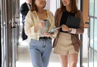 Two women talking and walking through a bright corridor with glass-partitioned offices.