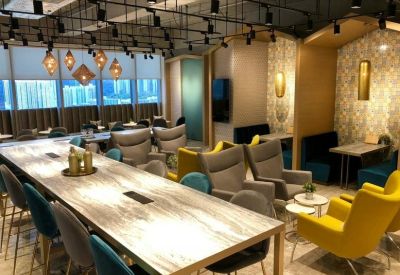 Spacious communal lounge with a long marble-patterned table, grey armchairs, and yellow accent seating.
