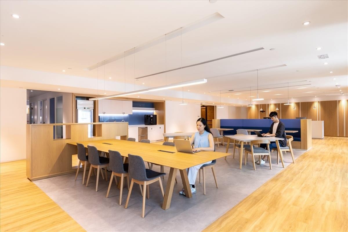 Vibrant coworking lounge with warm wood flooring, long communal tables, and comfortable seating.